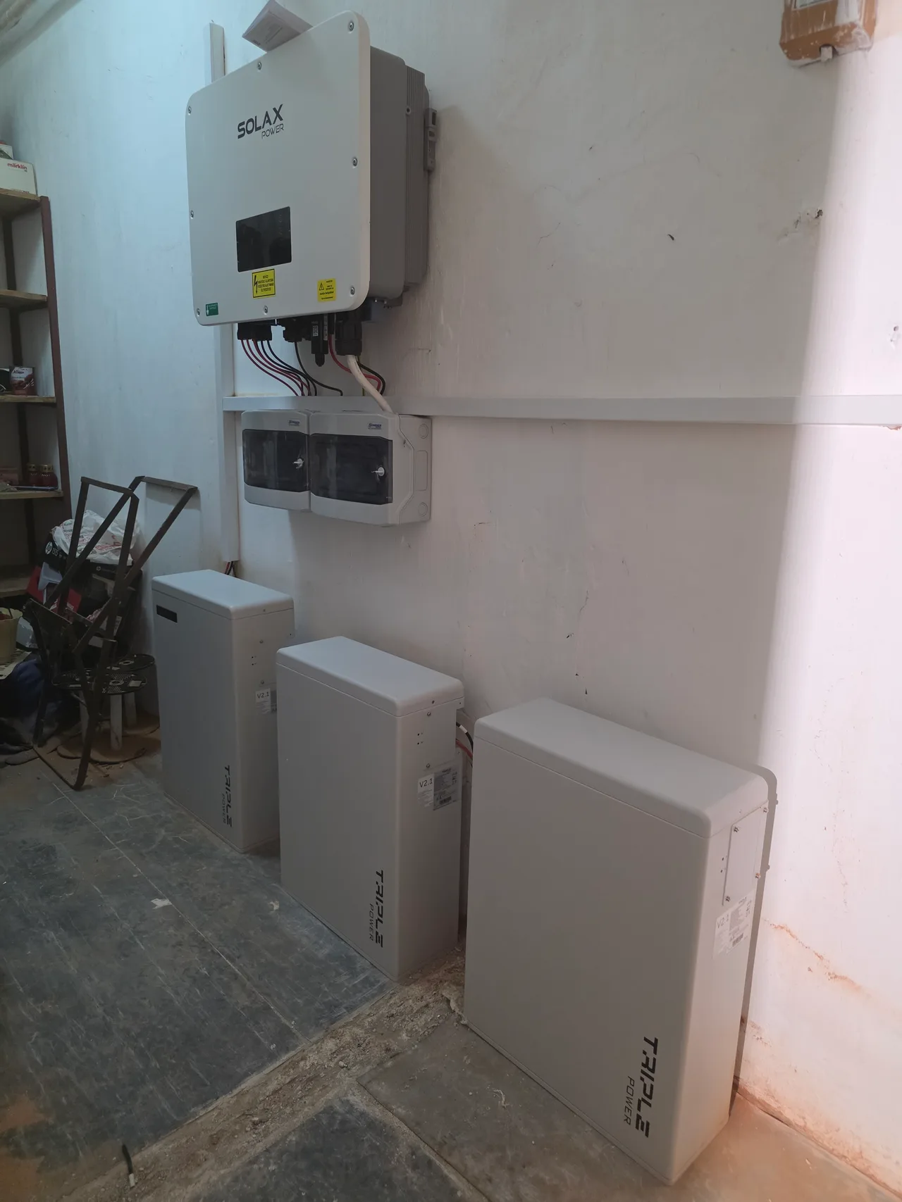 Falra szerelt SolaX Power inverter, alatta két kismegszakító-szekrény, a padlón három Triple Power akkumulátor-modul.